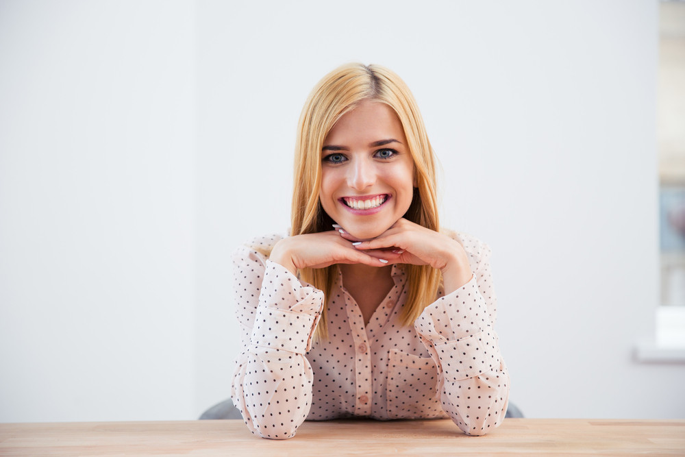 Smiling blonde businesswoman sitting at the desk over gray background and looking at camera